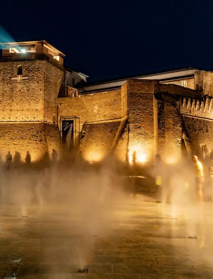 Château illuminé la nuit avec des effets de brouillard et des gens en mouvement.
