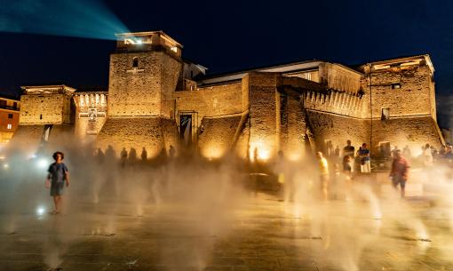 Château illuminé la nuit avec des effets de brouillard et des gens en mouvement.