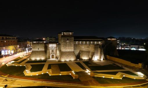 Castello medievale illuminato di notte con giardini moderni e strade circostanti.